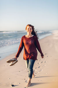 Smiling young woman walking at beach against clear sky during sunset