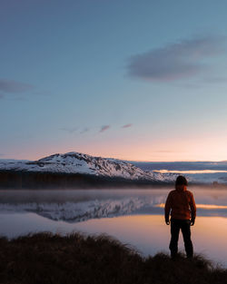 Rear view of man standing by lake against sky during sunset