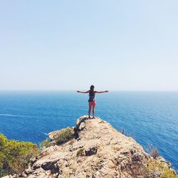 Rear view of woman standing on cliff while looking at sea against clear sky