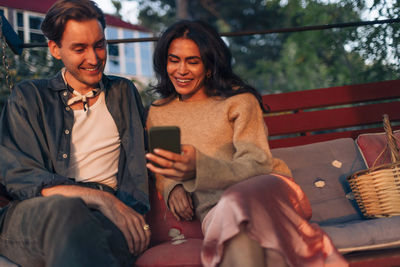 Smiling woman sharing smart phone with male friend while sitting on bench in back yard