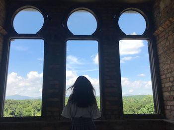 Rear view of woman standing against sky seen through window