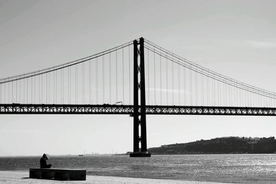 Woman sitting by 25 de abril bridge over tagus river against sky