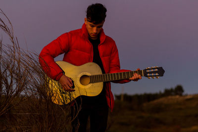 Young man playing guitar against sky at dusk