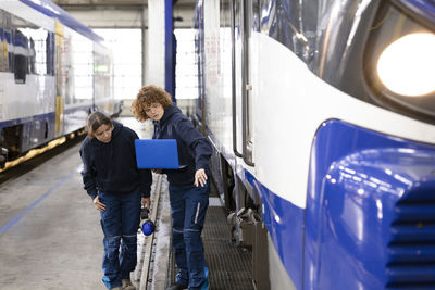 Engineer holding laptop explaining to trainee about monorail in factory