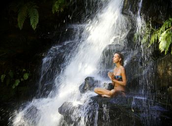 Woman in bikini meditating by waterfall on rock formation