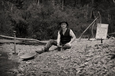 Portrait of man sitting on riverbank using mortar and pestle by signboard