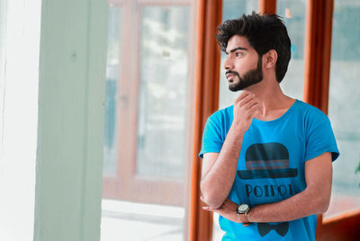 Young man looking away while standing against window