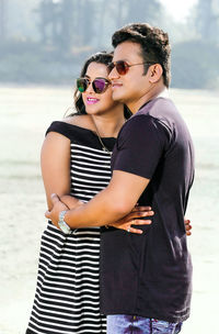 Couple wearing sunglasses while standing with arms around at beach