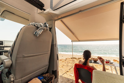 Rear view of woman sitting by motor home against sea