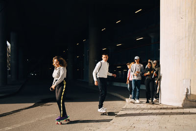 Friends looking at man and woman skateboarding on road during sunny day