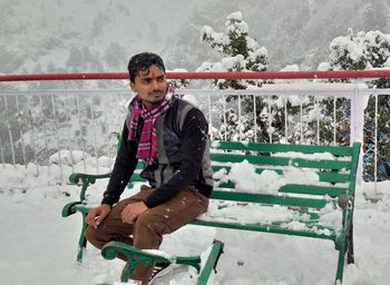 Young man looking away while sitting on bench in snow