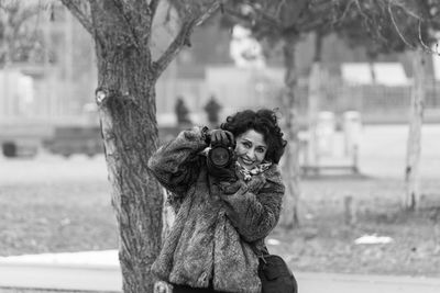 Portrait of smiling woman with dslr standing against tree at park