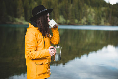 Side view of stylish female hipster wearing a hat, holding and drinking coffee from moka by the lake
