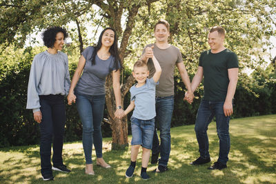 Portrait of playful son holding hands with smiling homosexual parents in backyard