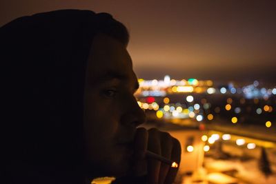 Close-up of young man smoking cigarette against illuminated city at night