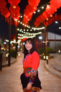 Portrait of smiling young woman standing against illuminated lights at night