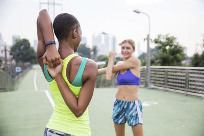 Woman in sports clothing stretching with friend on footpath