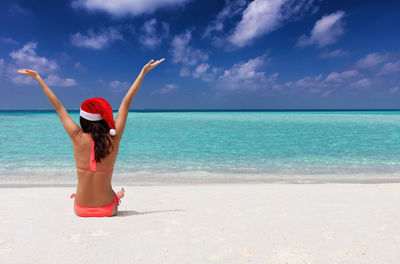 Rear view of woman wearing bikini and santa hat sitting at beach against sky