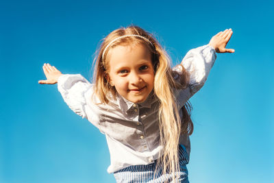 Portrait of smiling girl with arms raised standing against clear blue sky