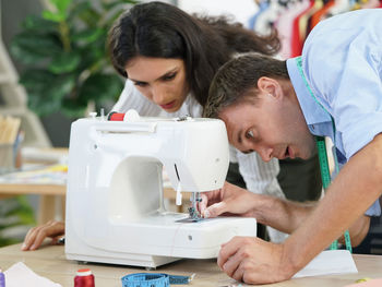Woman looking at man putting thread in sewing machine