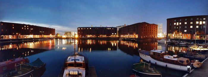 Sailboats moored on river by buildings against sky at dusk