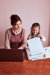 Smiling girl looking at paper on table while woman working on laptop