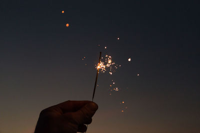 Cropped hand holding lit sparkler against sky at dusk
