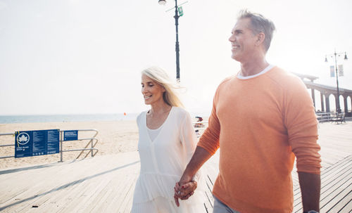 Smiling couple holding hands while walking at promenade by beach