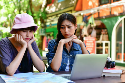 Sad young female friends with laptop and map on table at sidewalk cafe