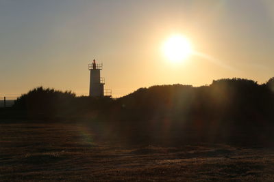 Lighthouse on field by buildings against sky during sunset