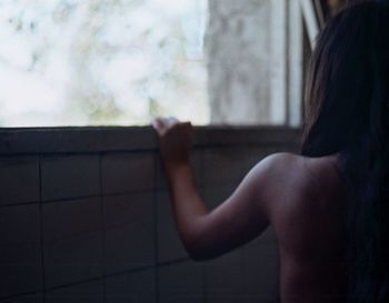 Rear view of shirtless woman standing by window