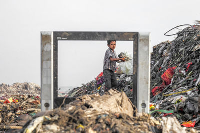 Boy standing by pile of garbage seen through metal against sky