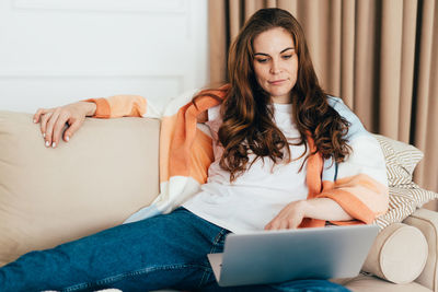 Young concentrated business woman lying on sofa at home uses laptop to watch movie.