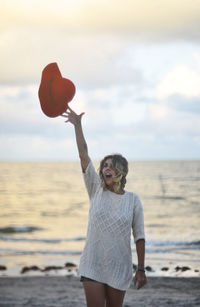 Woman throwing hat while standing at beach against sky