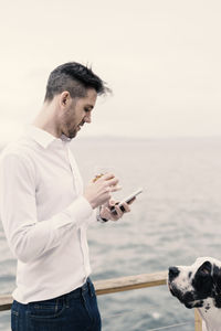 Young man using mobile phone while standing by dog on boat deck against sea