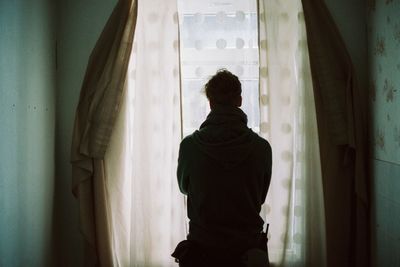 Rear view of man looking through curtained window