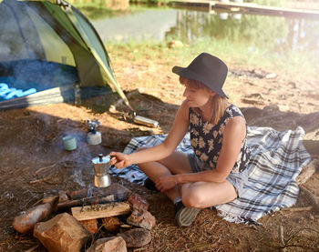 Young woman holding coffee pot over bonfire by tent during vacation