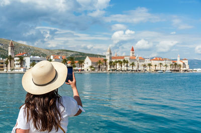 Rear view of young woman using mobile phone, taking photos of beautiful cityscape of trogir, croatia