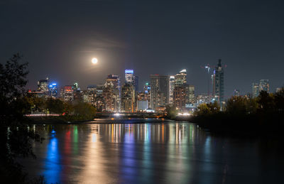 Illuminated buildings by river against sky at night