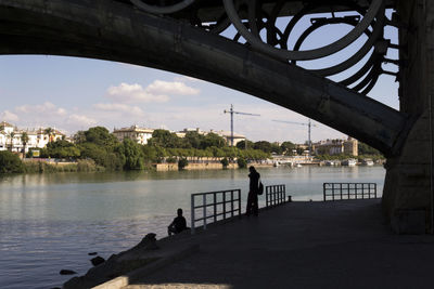 Silhouette person under bridge by river in city