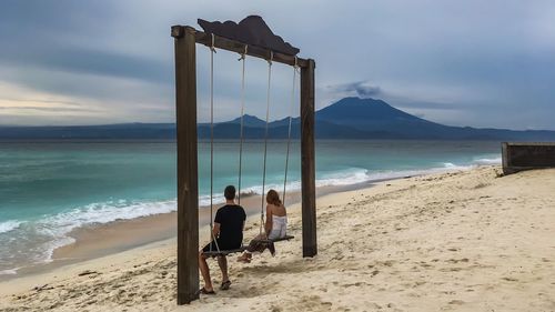 Couple sitting on swings at beach against cloudy sky