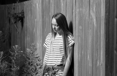 Sad teenage girl looking at plants while standing by wooden fence at backyard