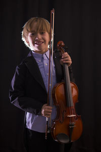 Boy holding violin while standing against black background