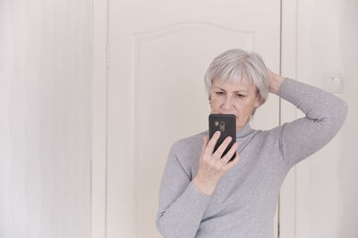 A gray-haired senior woman in a gray turtleneck looking attentively at the smartphone at home. 