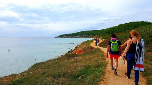 People walking on trail by sea at minorca against cloudy sky