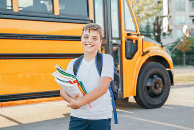 Portrait of smiling boy holding books standing by school bus