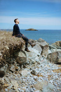 Man sitting on rock by sea against sky