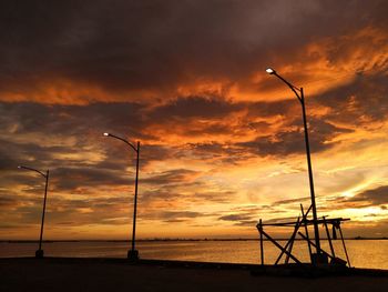 Silhouette street lights by sea against orange sky