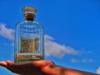 Close-up of hand holding glass bottle against blue sky