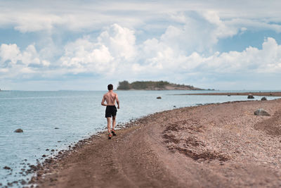 Rear view of shirtless man running at beach against cloudy sky